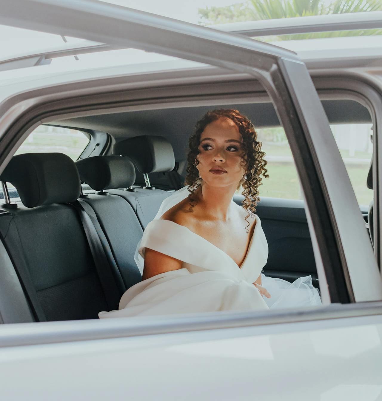 A bride in a white wedding gown sitting inside a luxury car, looking outside with an elegant expression.