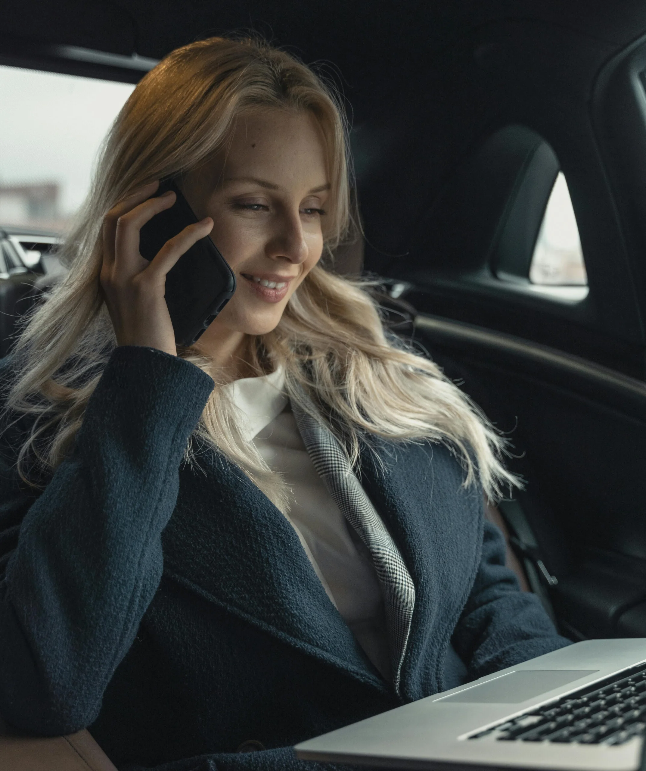 A professional woman with blonde hair is sitting in the back of a luxury car, engaged in a phone call while working on her laptop. The car's elegant interior provides a comfortable and private environment.