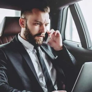 Business professional in a luxury car, talking on the phone while working on a laptop.