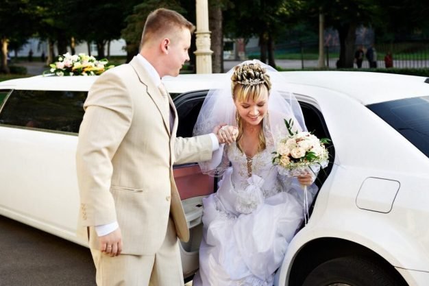 Groom helping his bride out of a luxury white limousine, holding her hand on their wedding day.
