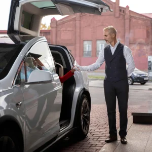 A professional chauffeur assisting a passenger into a luxury car with upward-opening doors.